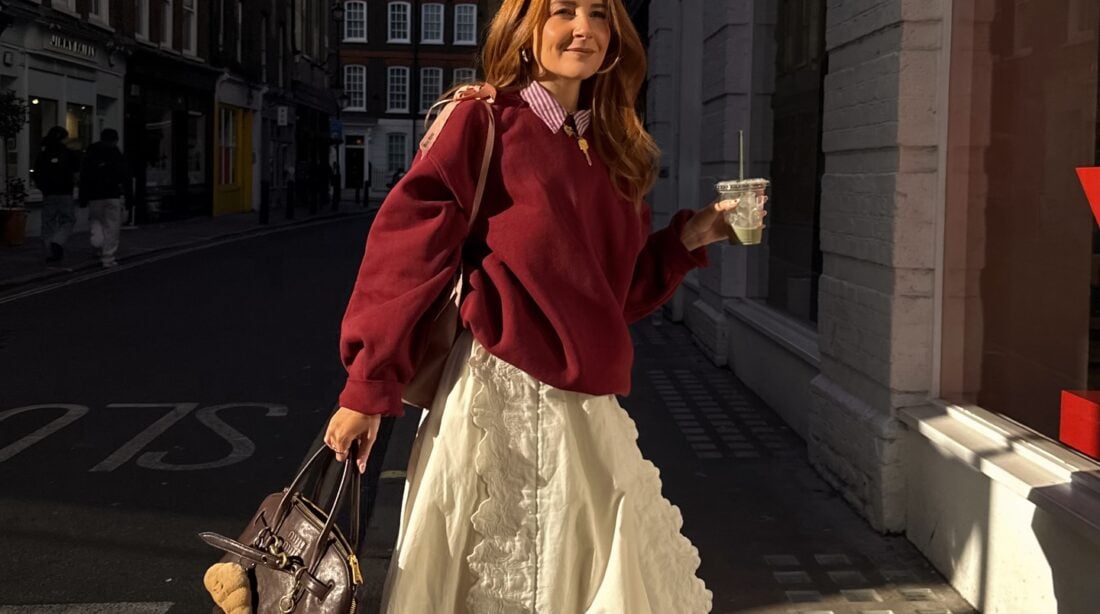 Mulher jovem, magra e de pele clara com cabelos ruivos. Ela usa look com suéter e saia