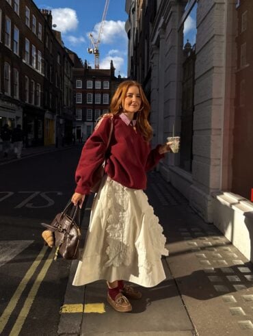 Mulher jovem, magra e de pele clara com cabelos ruivos. Ela usa look com suéter e saia
