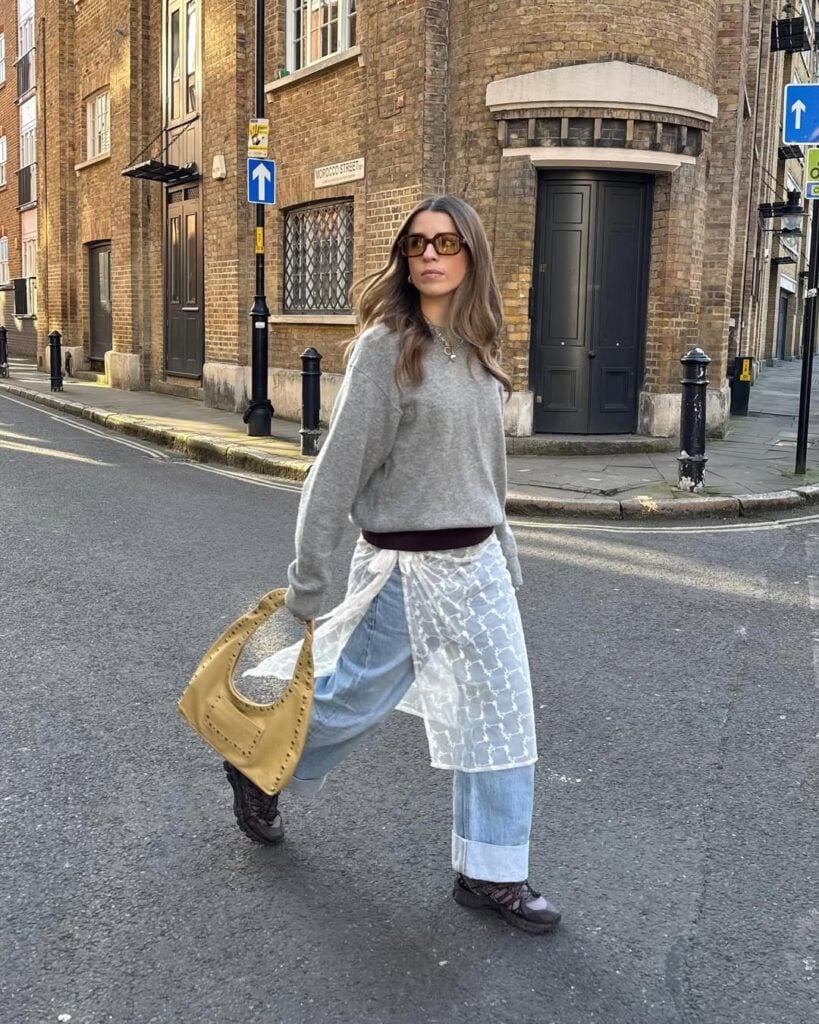 Mulher jovem de pele clara e cabelo castanho. Ela usa look com suéter cinza, calça jeans clara, saia com detalhes de renda branca, tênis, óculos de sol quadrado e bolsa